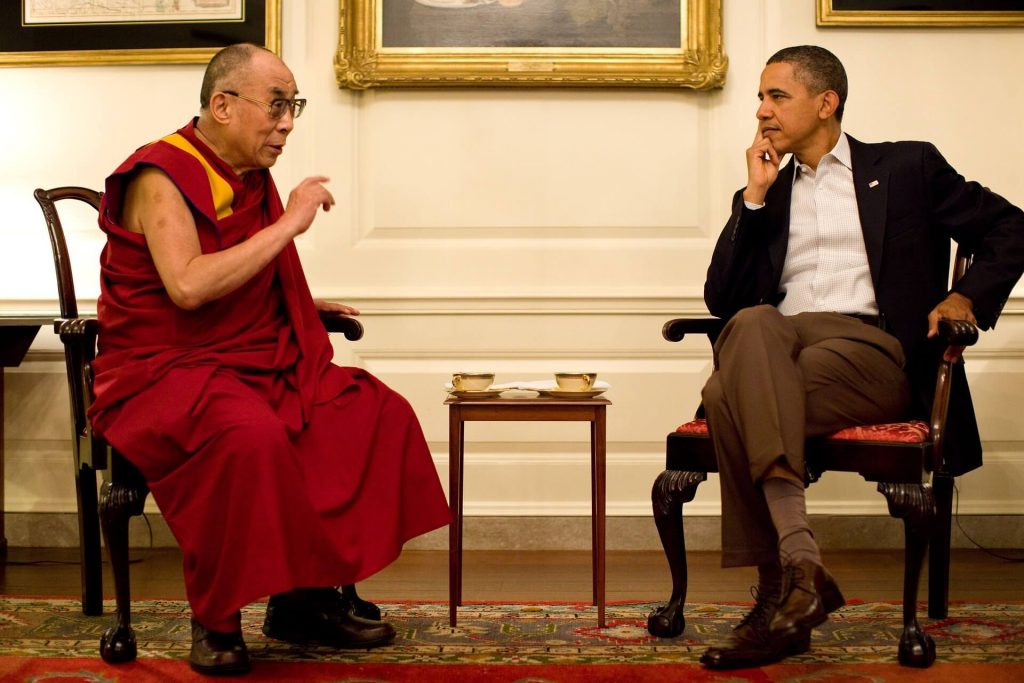 Barack Obama und der Dalai Lama im Gespräch miteinander. Obama sitzt mit überschlagenen Beinen und hört zu während der Dalai Lama lebendig und gestenreich spricht.